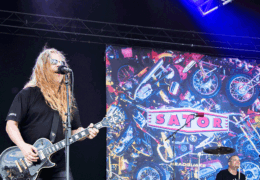 Chips Kiesbye sjunger i en mikrofon. I bakgrunden en skärm med bandnamnet "Sator".