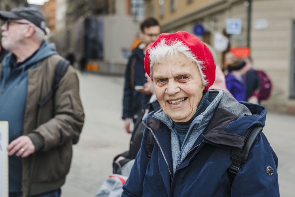 En kvinna i 80-årsåldern med knallröd basker och kort grått hår ler och tittar in i kameran.
