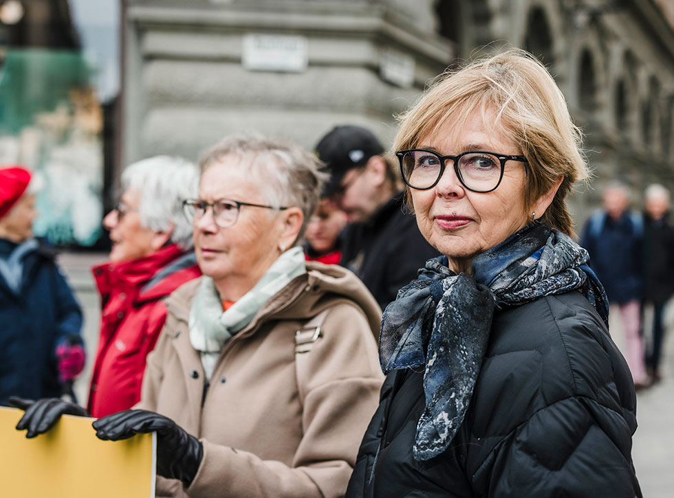 En äldre kvinna med kort grått hår i beige jacka håller händerna på ett plakat. Bredvid henne står en kvinna i svart jacka och glasögon som tittar rakt in i kameran.