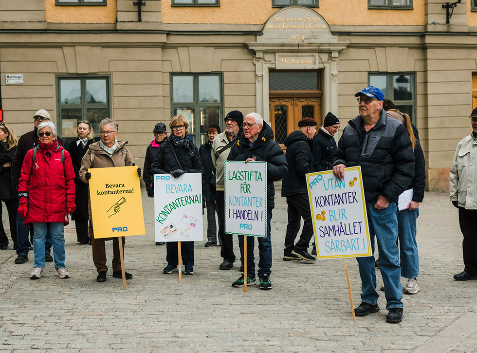 En grupp pensionärer står på ett torg med skyltar framför sig med budskap om kontanternas bevarande.