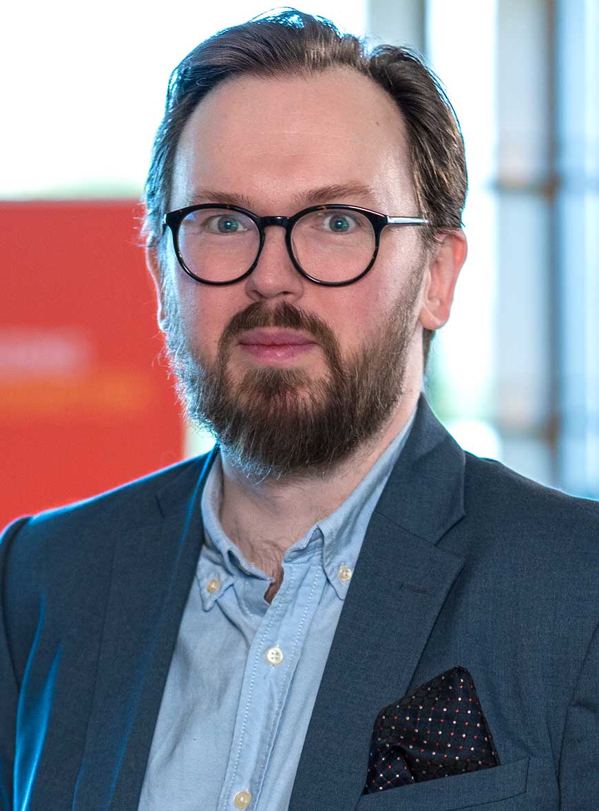En man med kammat hår, svartbågade glasögon och skägg iförd mörk kavaj och skjorta.